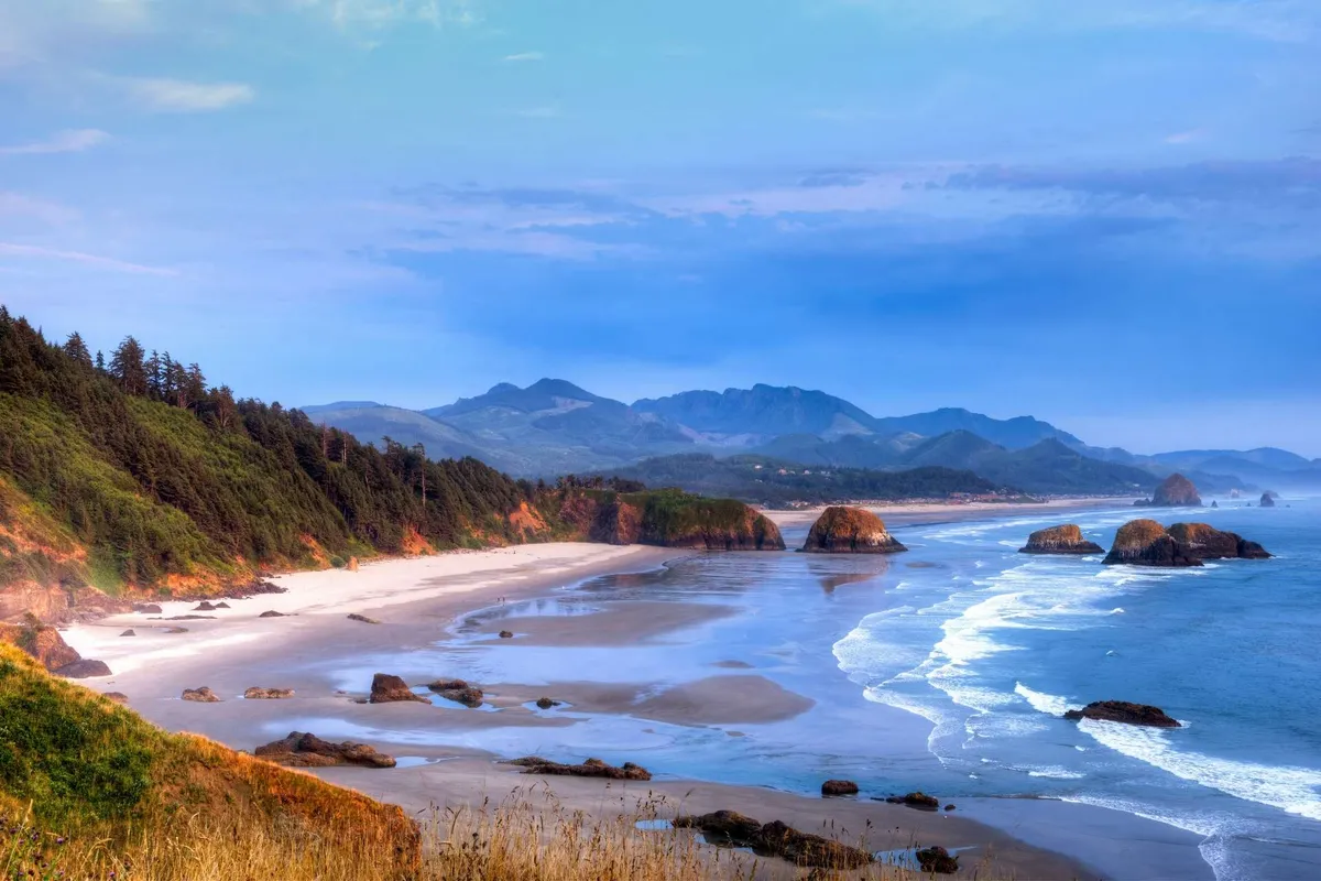 A panoramic view of the expansive beach, with ocean waves rolling in, striking rock formations dotting the shoreline, and distant mountains creating a dramatic backdrop. The tranquil setting is perfect for exploring, beachcombing, and family fun.