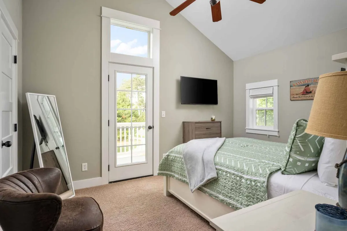 This bedroom boasts a queen bed with a green patterned quilt, a large window letting in plenty of natural light, a TV mounted on the wall, and a mirror reflecting sunlight. The room feels open, comfortable, and ready for relaxation.