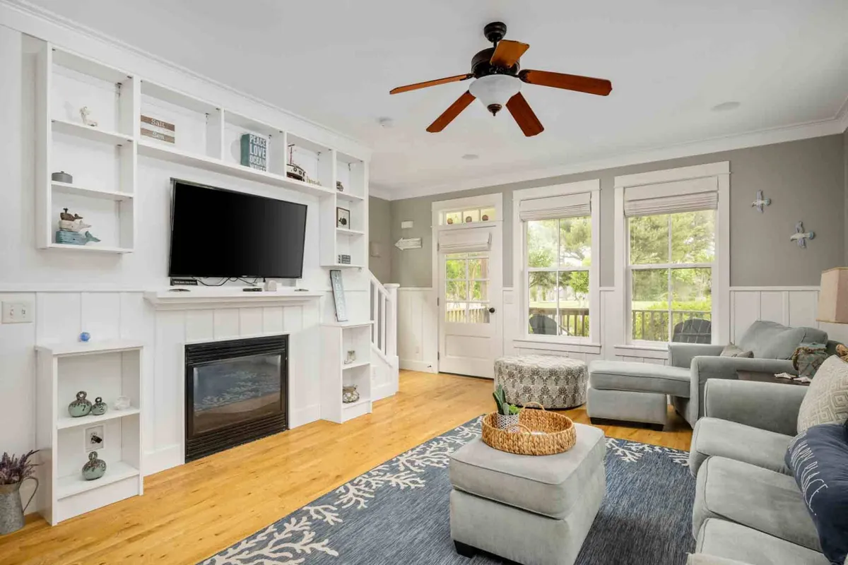 A cozy living room with a fireplace, large windows bathing the area in sunlight, a comfortable seating arrangement, and decorative shelves. The fireplace serves as a focal point, echoing the warmth and inviting decor described.