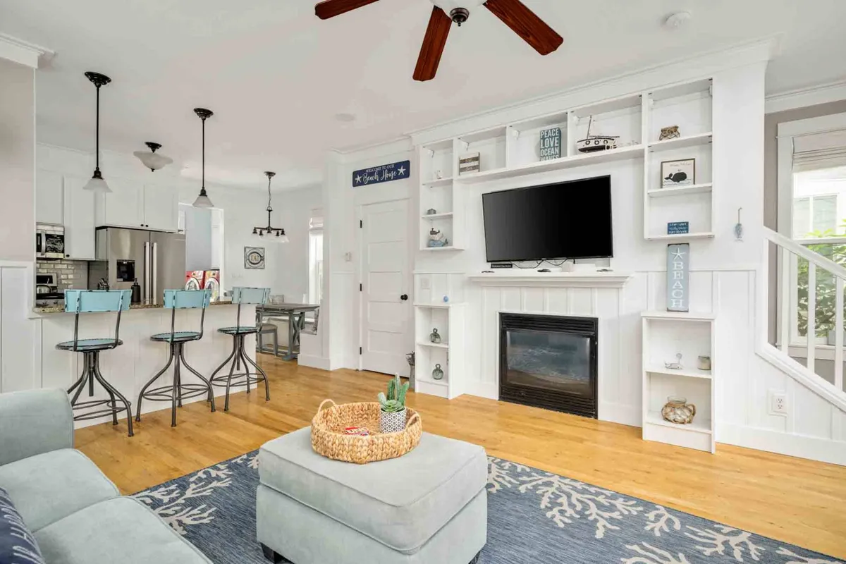 The image showcases a warm and inviting open-concept living area with a plush sofa facing a glowing fireplace and entertainment center, complemented by cozy bar stools at the kitchen counter. The space exudes comfort, perfect for relaxing, family gatherings, or enjoying quiet mornings with a cup of coffee.
