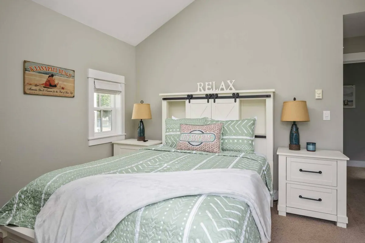 The image features a serene bedroom with a bed adorned with a 'RELAX' sign, decorative pillows, and lamps on nightstands. Beach-themed wall art and soft, natural lighting contribute to a peaceful, coastal atmosphere.