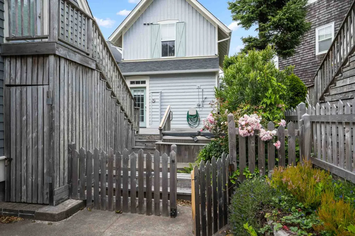 A welcoming exterior view of the house, featuring a wooden fence, flowering plants, garden stairs leading toward the upper level, and the inviting façade of the cottage. The lush landscaping and outdoor space highlight the transition from the peaceful home to the lively beach scene just beyond.