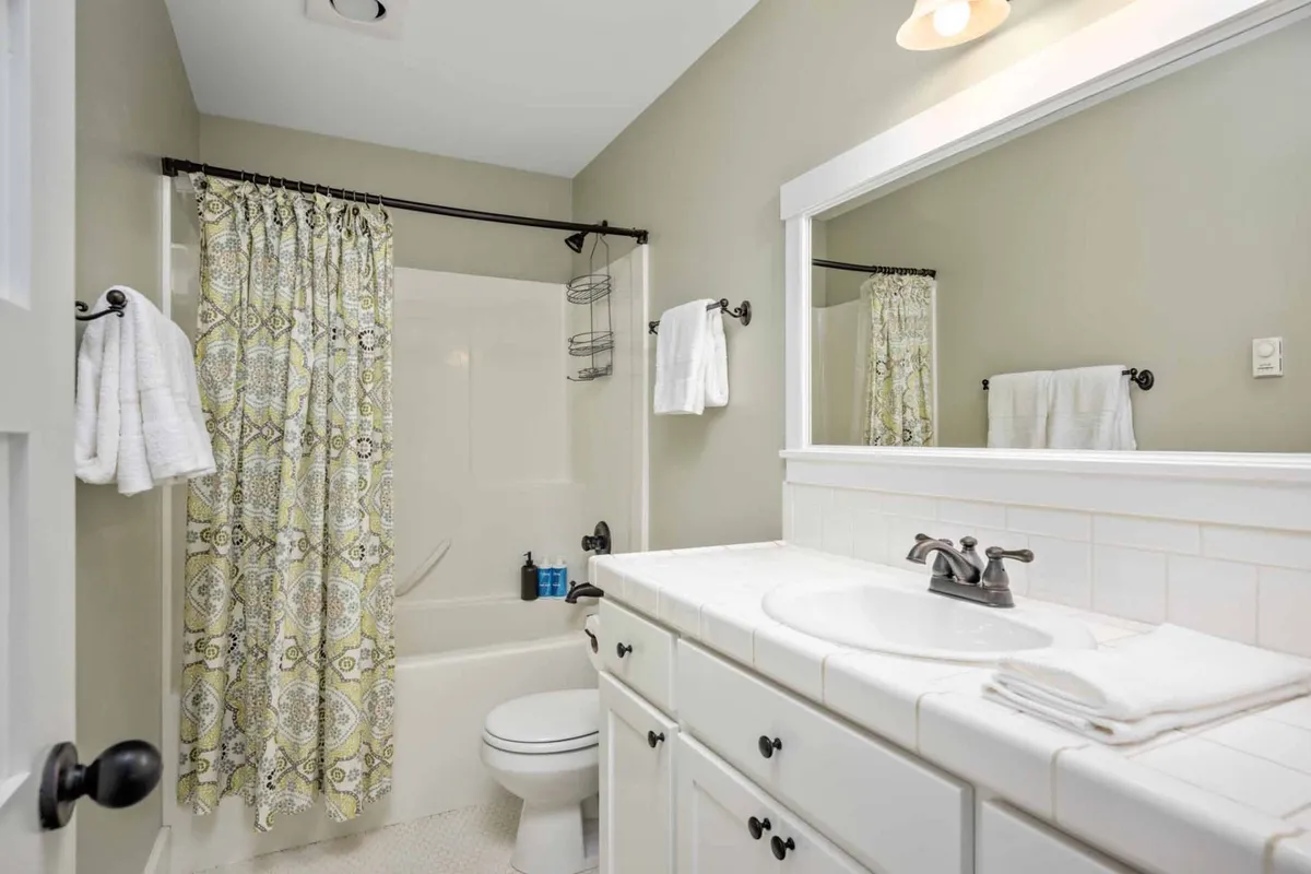 A bright, inviting bathroom with white cabinetry, a decorative shower curtain, and a mirror. The space feels clean and comfortable, ideal for refreshing after a day at the beach or pool.
