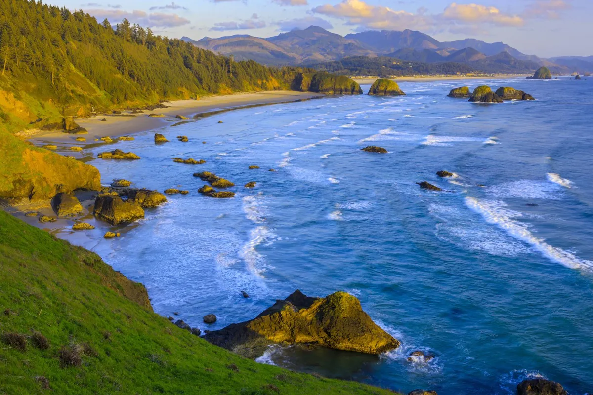 A sweeping view of the rugged coastline with crashing waves, dramatic rock formations, distant mountains, and lush greenery. The scene is tranquil and expansive, offering a sense of open air and natural beauty that epitomizes the Oregon coast.