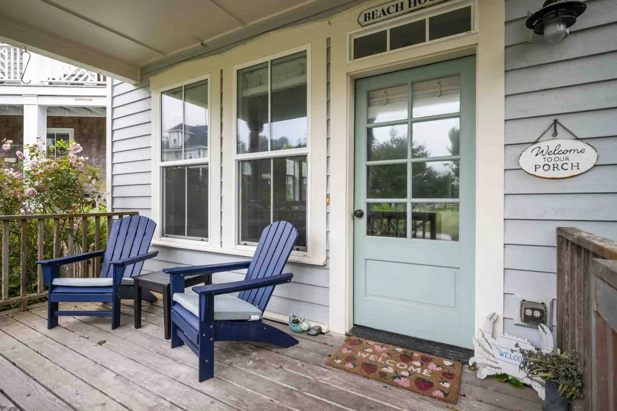The image showcases a charming, partially covered porch with blue Adirondack chairs and a welcoming mat, centered around a cheerful light blue door. The scene feels inviting, ideal for lingering over morning coffee or greeting friends and family.