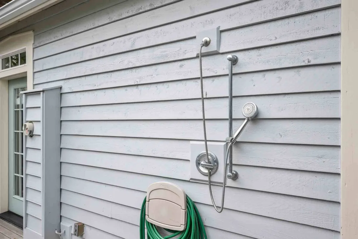 The image features an outdoor shower and a green hose against charming wooden siding, suggesting practical amenities for post-swim rinsing and gardening. This refreshing, functional space is set in a sunny outdoor environment, aligning with community leisure activities.