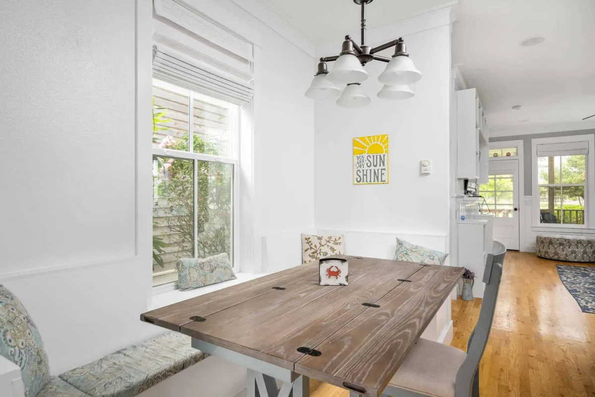 A cozy dining area featuring a wooden table surrounded by chairs, bathed in natural light from a window that offers a glimpse of lush greenery outside. The space is accented by a distinctive sunshine-themed wall art and a charming overhead light fixture, creating an inviting place for meals or morning coffee.