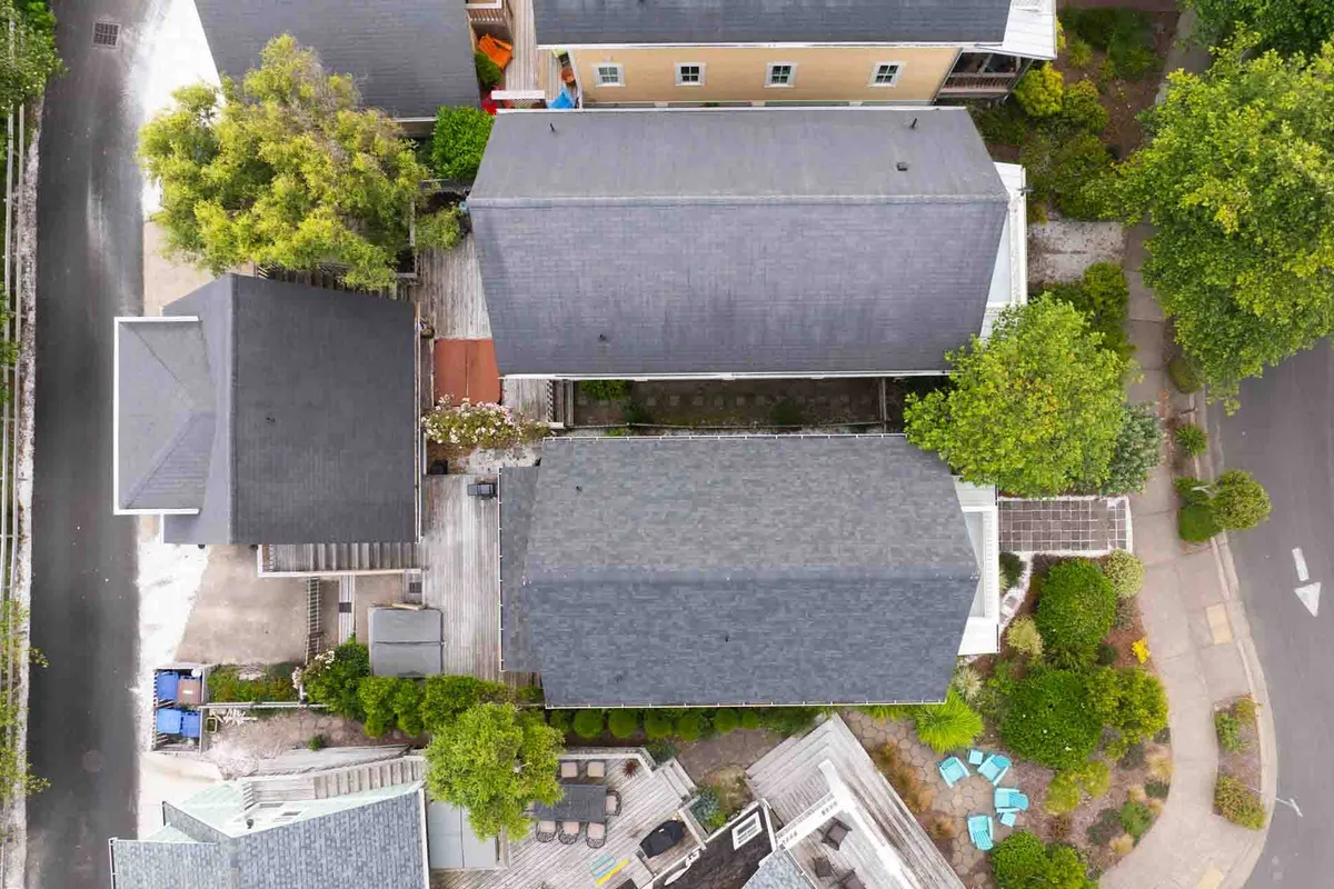 This image showcases the rooftops of several cottages surrounded by lush green trees, with a patio area and a walkway. People are seen engaging in outdoor activities, such as social gatherings and relaxing, while children play in the yard. The setting exudes a peaceful yet social atmosphere and highlights the community's shared outdoor spaces.
